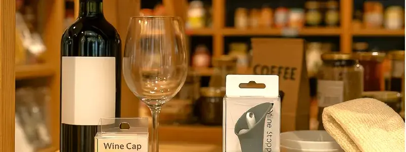 カルディのような雑貨店の店内写真。木製の棚にワインボトルやグラス、コーヒー豆、調味料瓶などが並び、柔らかい光に包まれた温かみのある雰囲気。ワインストッパーなどワイン関連の小物が置かれた構図