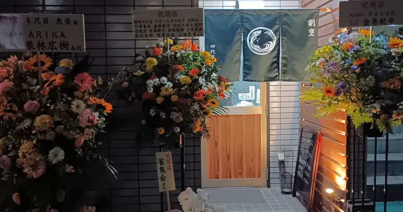 四代目魚盛の外観。開店祝いの花に囲まれた夜の入口と暖簾